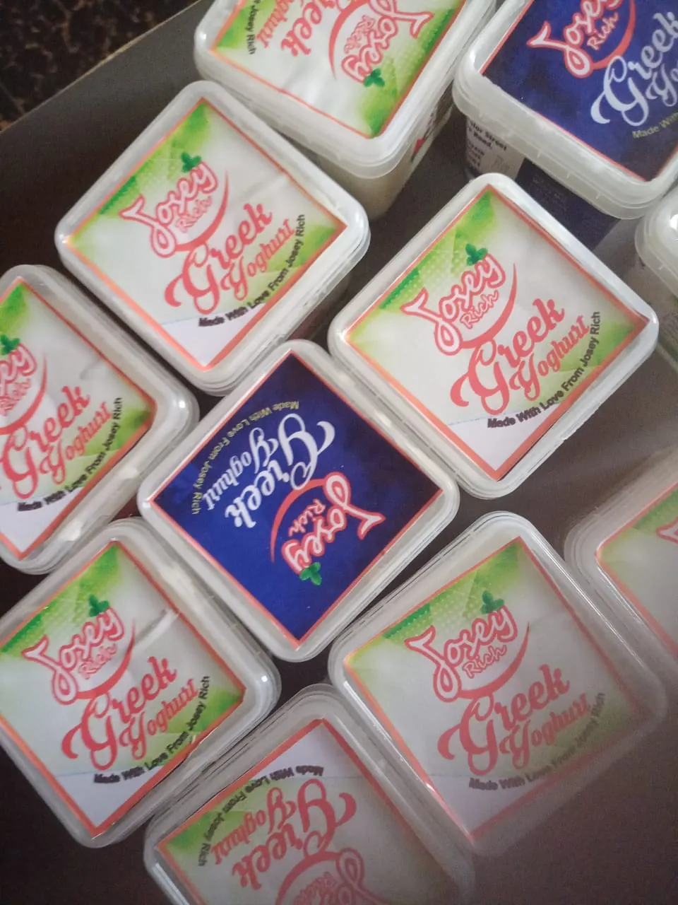 A tray of square Josey Rich Greek Yoghurt tubs seen from above, alternating white-and-green and navy lids, each printed with the coral script wordmark.