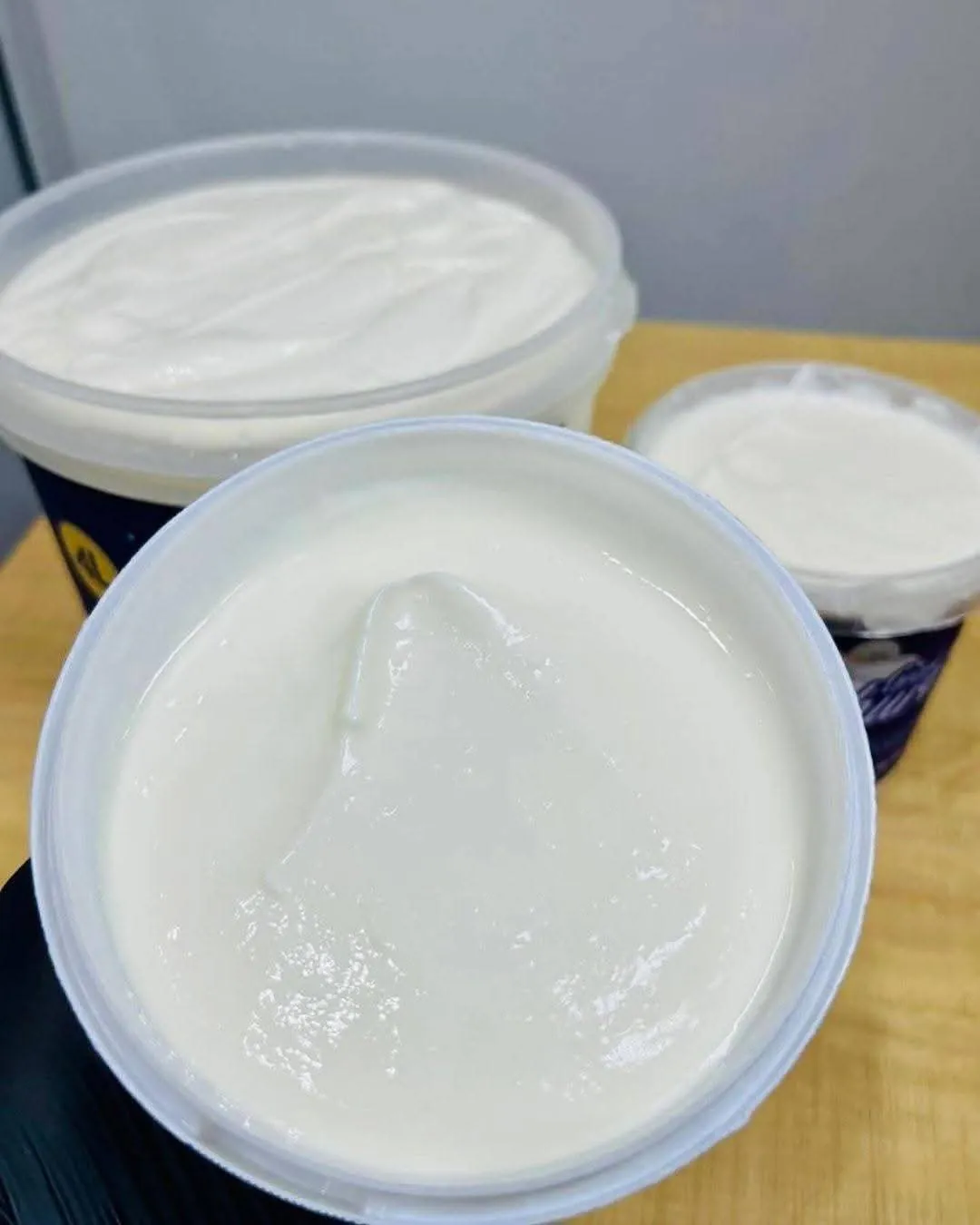 Three open tubs of Josey Rich Greek Yoghurt on a wooden table, thick and glossy white, the nearest one drawn up into a soft peak that holds its shape.