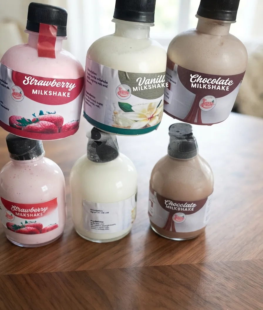 Six Josey Rich milkshake bottles on a dark wooden table — pink strawberry, cream vanilla and brown chocolate, each labelled with its flavour.