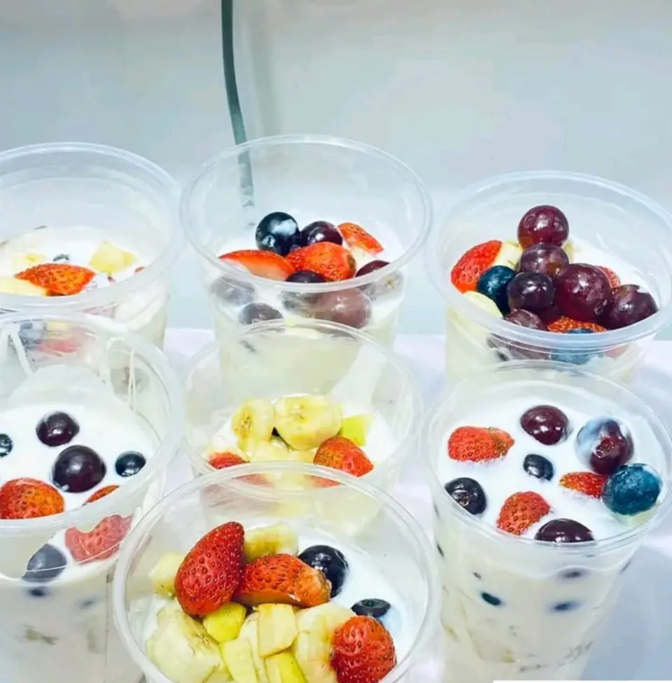 Seven clear cups of yoghurt parfait on a white cloth, each topped with sliced strawberry, banana, dark grapes and blueberries.