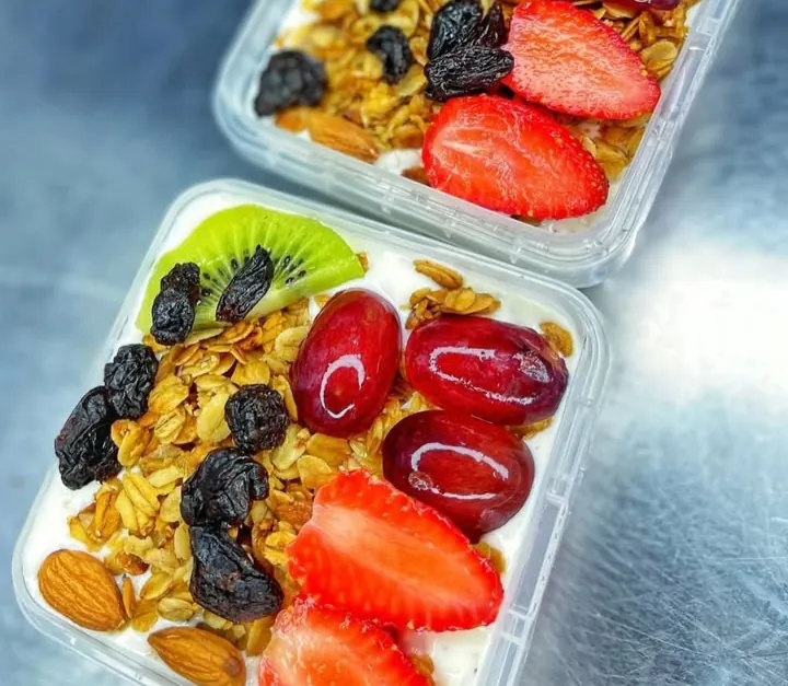Two clear trays of yoghurt topped with granola, almonds, raisins, red grapes, kiwi and halved strawberries, on a steel counter.